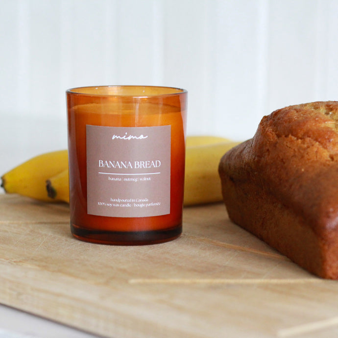 Banana bread loaf and 'Banana Bread' scented candle on a wooden cutting board with a white background